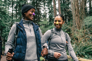 Two women with hiking equipment walking together during the day in a forest while using NÖZ sunscreen in their noses.